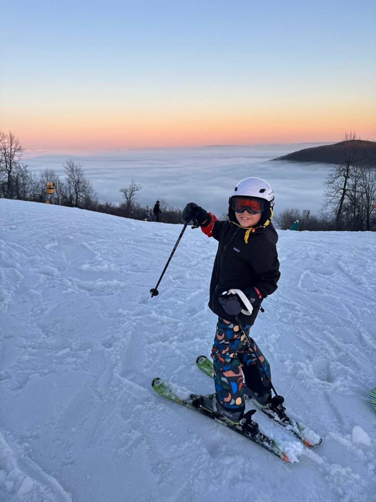 A Ski Club student pauses on a run at dusk, with a valley of low cloud behind the mountain.